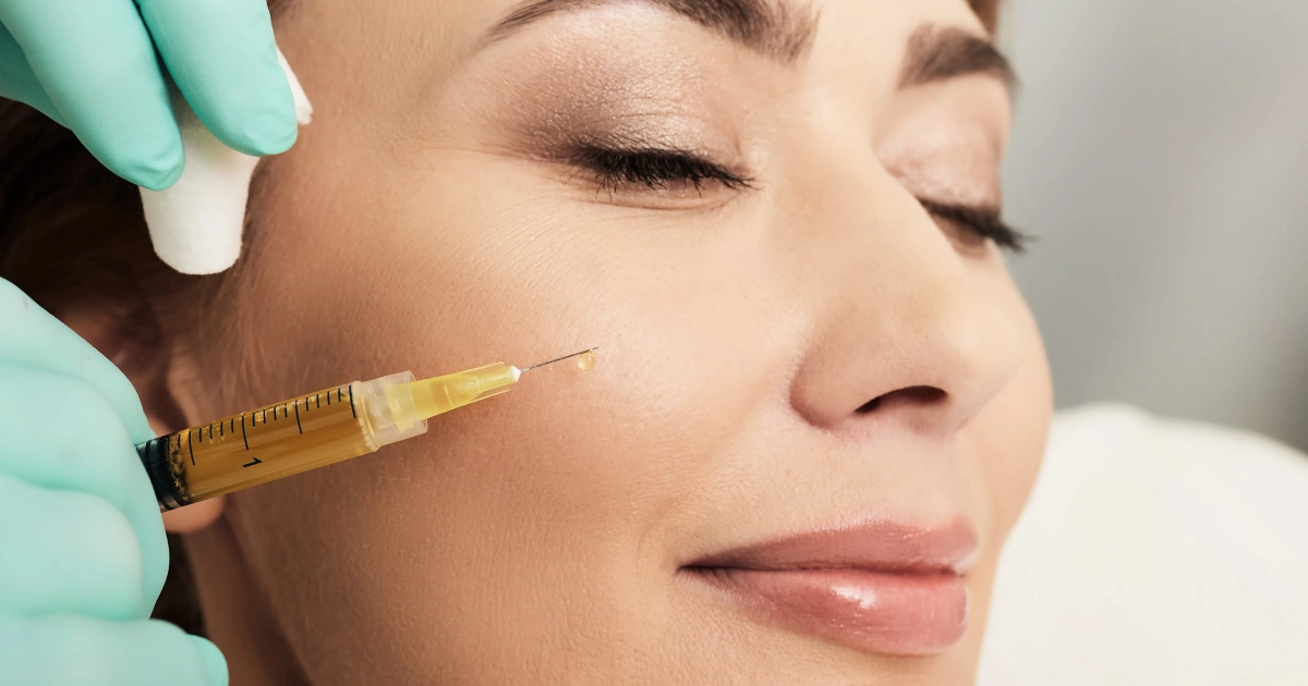 woman receiving a PRP facial rejuvenation in Olympia, with an injection near her cheekbone administered by a person in teal gloves holding a syringe of yellow fluid. The woman looks calm.